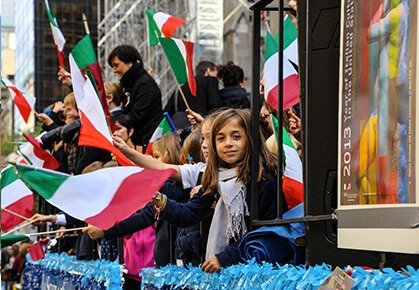 A young girl waves an Italian flag from a decorated parade float surrounded by people holding flags during a celebration.