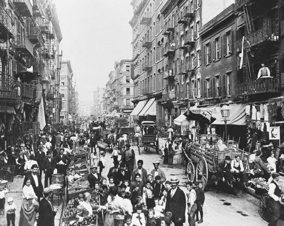 A bustling Italian-American marketplace in a 19th-century New York City street, filled with vendors, shoppers, and horse-drawn carts surrounded by tenement buildings.